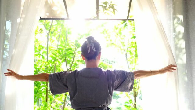 The woman was at the window in the bedroom. She opened the curtains on the window. In the morning and she looks at the view of mountains and trees at sunrise.