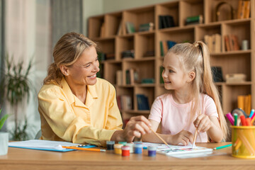 Pretty little girl painting with child specialist, sitting at table with female teacher and holding brush, copy space