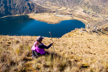 Latin woman, sitting on a hill, looking at a lagoon in the Peruvian Andes