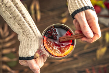 Hands of woman in a sweater are holding a glass cup with mulled wine on a wooden background with autumn leaves. Concept of a traditional warming beverage