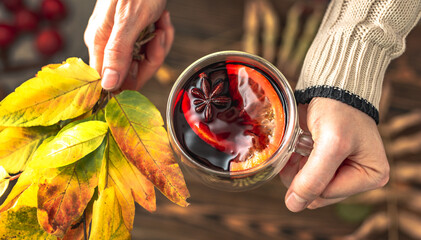 Hands of woman in a sweater are holding a glass cup with mulled wine and a bouquet of yellow autumn...