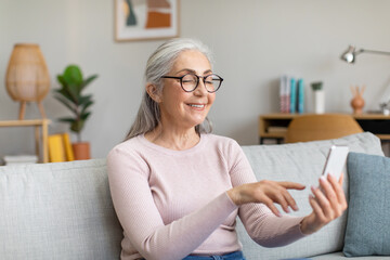 Obraz premium Smiling caucasian old gray-haired female in glasses have chat or video call, watch video on smartphone in room