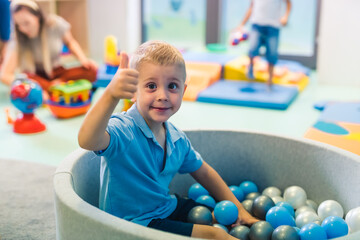 Happy toddler boy playing in a ball pit full of colorful balls, showing thumb up. Sensory play at the nursery school for kids wellness. High quality photo