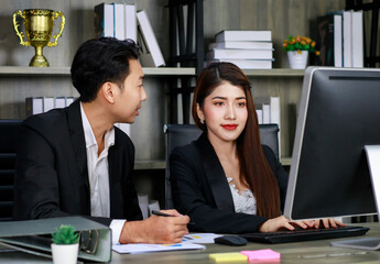 Millennial Asian young professional successful female businesswoman ceo entrepreneur in formal suit sitting smiling working via computer while male businessman secretary helping writing note on paper