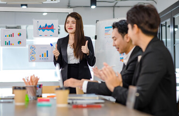 Asian young beautiful professional successful businesswoman presenter standing smiling in front of glass board after presenting while male female colleagues clapping hands thank you in meeting room
