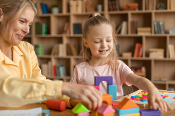 Friendly child development specialist observing girl preschooler, little child making pyramids from colorful wood blocks