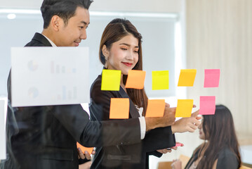 Asian young beautiful professional successful female businesswoman in formal suit standing smiling holding hand pointing presenting information data on sticky note on glass board to colleague