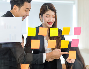 Asian young beautiful professional successful female businesswoman in formal suit standing smiling holding hand pointing presenting information data on sticky note on glass board to colleague