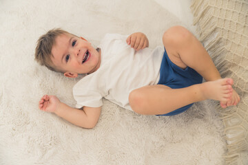 Amused caucasian preschooler boy in white t-shirt and dark-blue short lying on his back on the bed and laughing.Happy childhood concept. High quality photo - obrazy, fototapety, plakaty