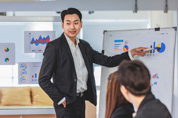 Asian handsome professional successful businessman presenter standing holding pen pointing presenting company graph chart paperwork document on glass board to male female colleagues in meeting room - obrazy, fototapety, plakaty