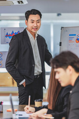 Asian handsome professional successful businessman presenter standing holding pen pointing presenting company graph chart paperwork document on glass board to male female colleagues in meeting room