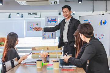 Asian handsome professional successful businessman presenter standing holding pen pointing presenting company graph chart paperwork document on glass board to male female colleagues in meeting room