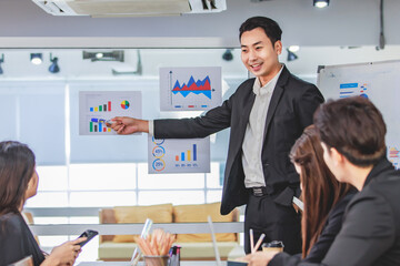 Asian handsome professional successful businessman presenter standing holding pen pointing presenting company graph chart paperwork document on glass board to male female colleagues in meeting room