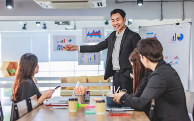 Asian handsome professional successful businessman presenter standing holding pen pointing presenting company graph chart paperwork document on glass board to male female colleagues in meeting room