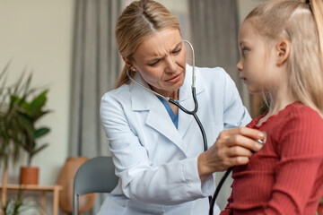 Worried pediatrician doctor examining little girl's heart beat and lungs with stethoscope, doing pediatric checkup