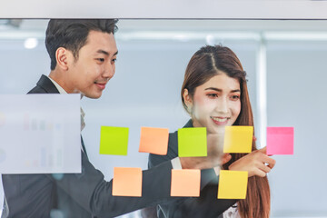 Asian young beautiful professional successful female businesswoman in formal suit standing smiling holding hand pointing presenting information data on sticky note on glass board to colleague