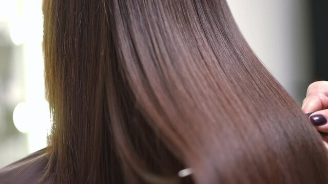 Close-up shaking long laminated hair with hair brush in slow motion. Brunette unrecognizable Caucasian young woman after keratin treatment in beauty parlor indoors with hairstylist