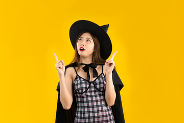 Halloween theme, young asian woman in black dress wearing witchcraft and hat posing finger pointing for you advertising on yellow background.