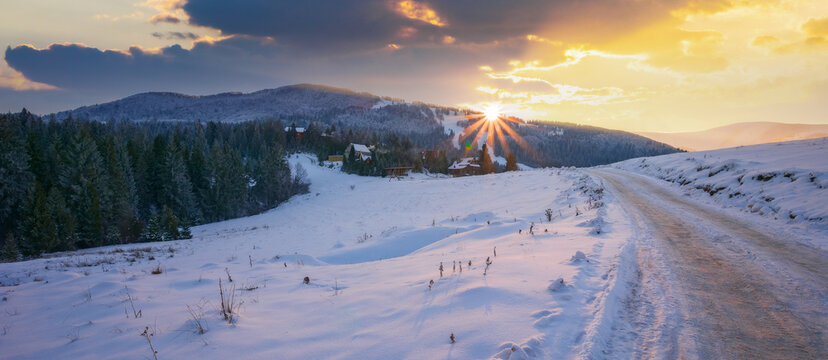 countryside landscape in mountains at sunset. beautiful scenery with coniferous forest on the snow covered hills in evening light. sun above the ridge. village in the distance