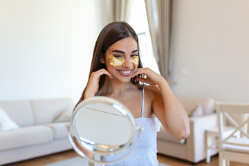 Smiling young woman applying eye hydration mask under her eyes. Female enjoy making face massage, peeling, put on mask under eyes in front of the mirror. Young woman looking in the mirror