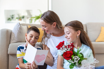 Young mother with a bouquet of roses laughs, hugging her son, and сheerful girl with a card congratulates mom during holiday celebration in kitchen at home