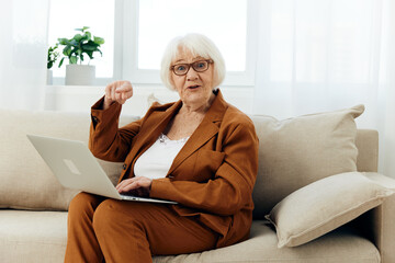 a joyful elderly woman is sitting on the sofa in a bright apartment in a brown suit, holding a laptop on her lap and pleasantly smiling with happiness showing a hand gesture as a sign of success