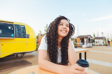 A beautiful cheerful girl with dreadlocks is sitting on a food court and drinking coffee. fast food cafe on the street. a woman drinks coffee in a cafe. autumn