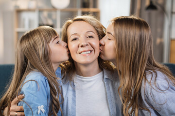Mother's Day. Caucasian mother holds her two children, daughters while they hug and kiss her. Happy family of mom and girls sisters sitting on sofa at home.