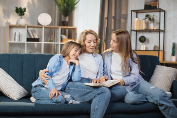 Happy loving family at home. Pretty young caucasian mother reading a book to her daughters sitting on couch in bright living room. Free time educative activity.