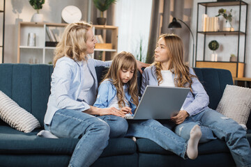 Happy family of mother and two cute daughters sitting together on comfortable couch, using laptop, resting and spend free time at home, recording video in living room.