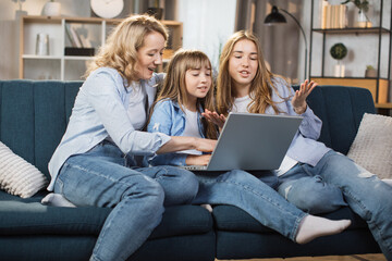 Happy family with two children having good time using laptop. Young smiling mother with cute blond daughters sitting on sofa at home looking at computer screen and watching video.