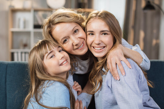 Portrait of happy two daughters hugging pleasant smiling middle age mother, relaxing together on couch. Affectionate two generations family looking at camera, posing for photo at home.