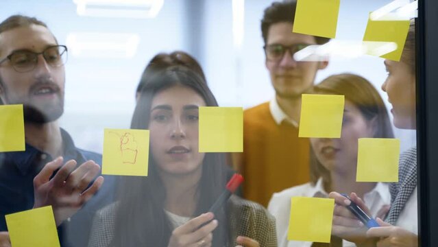 Business team brainstorm writing ideas on paper sticker notes and post it on glass wall. Startup coworkers discuss a strategy of work plan for product development at scrum meeting in corporate office.