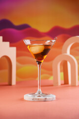 One martini glass with ice cube and brown alcoholic drink with black berry on bright colorful red and orange background