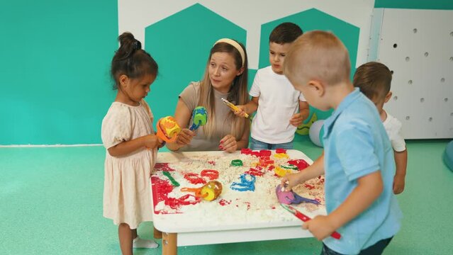 Fun sensory play with moldable kinetic sand at nursery school. Toddlers standing around the table and using different tools for sculpting sand such as colorful and textured rolling pins, cutters
