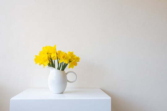 Daffodils in white jug on table against beige wall with copy space (selective focus)