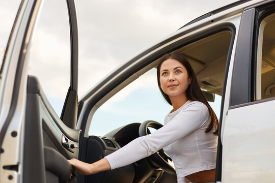 Image of beautiful brunette young adult woman with long hair getting out of a modern car looking in distance with calm confident expression, female traveling alone.
