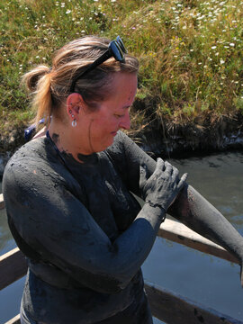 Blonde Caucasian woman smears therapeutic black mud on her arms and body. Her body is completely black, except for her face. Salted lakes of Ocna Sibiului.