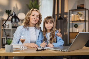 Portrait of joyful small daughter and her middle age mother doing homework and looking at camera. Young mom and her cute girl happy to make right tasks, sitting at home.