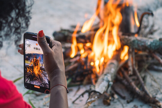 Girl taking photos or video with smartphone while preparing coffee to tea on a bonfire with branches and flames outdoor in the nature