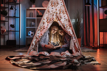 Caring caucasian older sister or nanny with her younger sister inside toy wigwam while reading fairytale. Dark atmosphere at cozy home. © Sofiia Shunkina