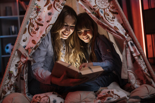 Caring caucasian older sister or nanny with her younger sister inside toy wigwam while reading fairytale. Dark atmosphere at cozy home.