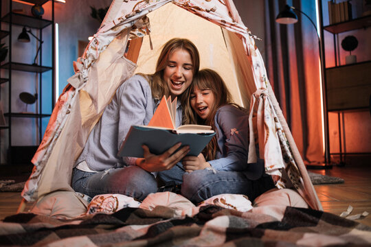 Caucasian pretty blond sisters sitting in wigwam and reading book during evening time. Cozy home and family concept.