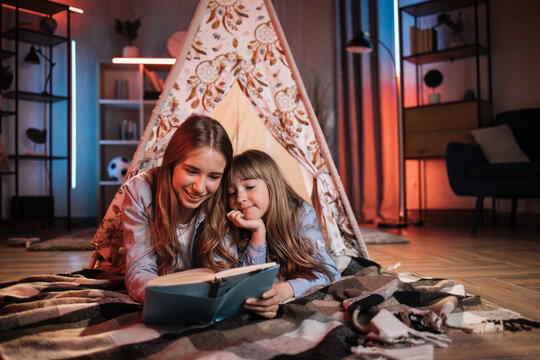 Happy caring nanny or older sister reading fairytale to her younger sister during evening time outside teepee tent lying on the floor at dark cozy room.