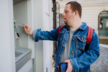 Happy young man with Down sydrome using a street ATM machine and withdrawing money.