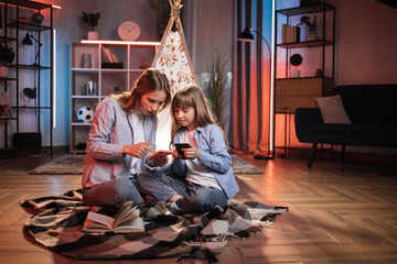 Fototapeta na wymiar Caring caucasian older sister or nanny with her younger sister outside toy wigwam using their smart phones. Dark atmosphere at cozy home.