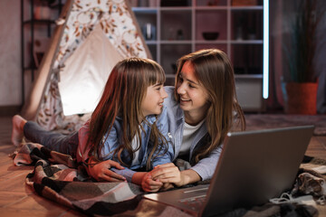 Caucasian pretty blond sisters lying on floor outside wigwam while using laptop for watching movie during evening leisure time. Cozy home and family concept.