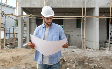 Young engineer in a construction helmet looking at real estate projects floor plan. The construction site has scaffolding made of wood.