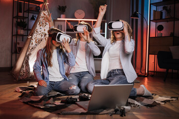 Happy mother and cute daughters sitting near toy wigwam at home and using modern VR headset during evening time. Caucasian family using modern technologies for playing at home.