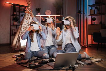 Happy mother and cute daughters sitting near toy wigwam at home and using modern VR headset during evening time. Caucasian family using modern technologies for playing at home.